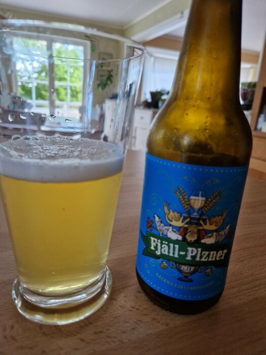 Ölglas halvfyllt, flaska etikett "Fjäll-Pilsner", inomhus, köksbakgrund, dagsljus genom fönster.