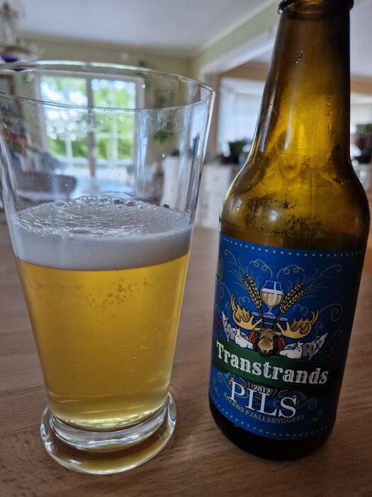 Ett glas med ljus öl och en ölflaska med etikett "Transtrands Pils". Inomhus, bakgrund med heminredning.