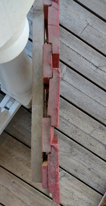 Rödmålade tegelstenar staplade ovanpå varandra på ett trägolv, nära en vit pelare.