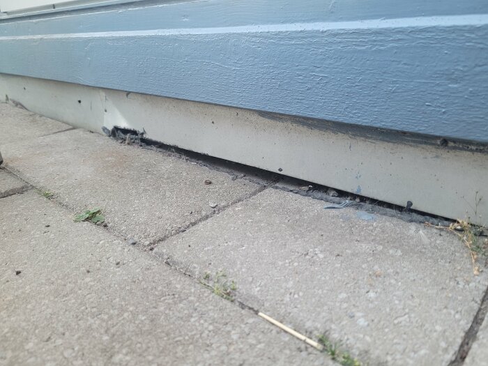Blåmålad byggnadsvägg möter trottoar; skadad del och mindre skräp vid byggnadens grund.