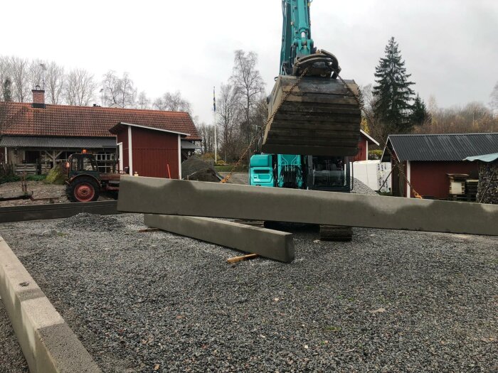 Byggplats med grävmaskin, betongelement och grus. Lantlig miljö, molnig himmel, röda byggnader och svensk flagga i bakgrunden.