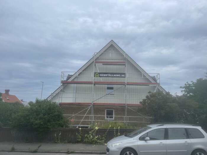 Ett hus med byggnadsställningar, molnig himmel, parkerad silverfärgad bil framför.