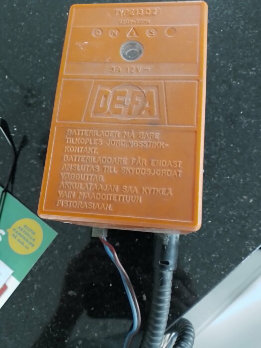 Orange batteriladdare på köksbänk, kablar anslutna, text på svenska, instruktionslapp i bakgrunden.