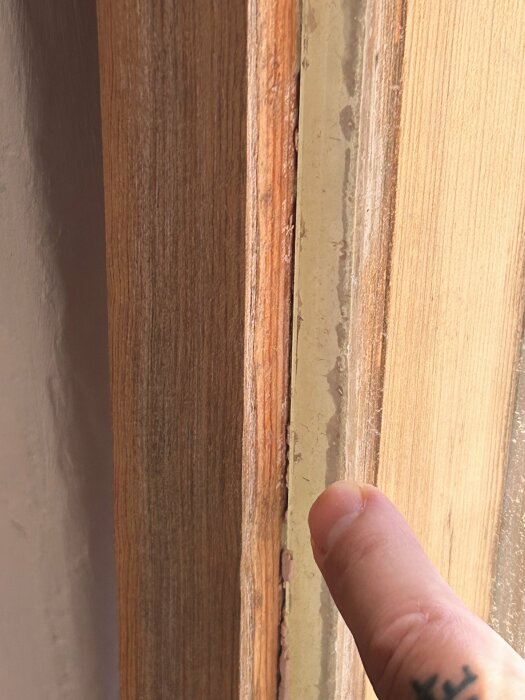 Ett finger pekar på en skada vid en dörrkarm; sliten färg och trä syns.