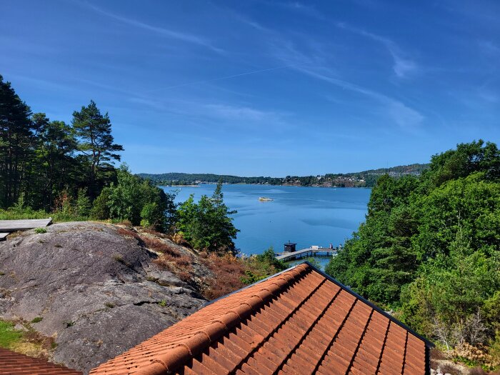 Skärgårdsutsikt, soligt, klart blått vatten, gröna träd, tegeltak, båt, brygga, avkopplande landskap, blå himmel, sommardag.