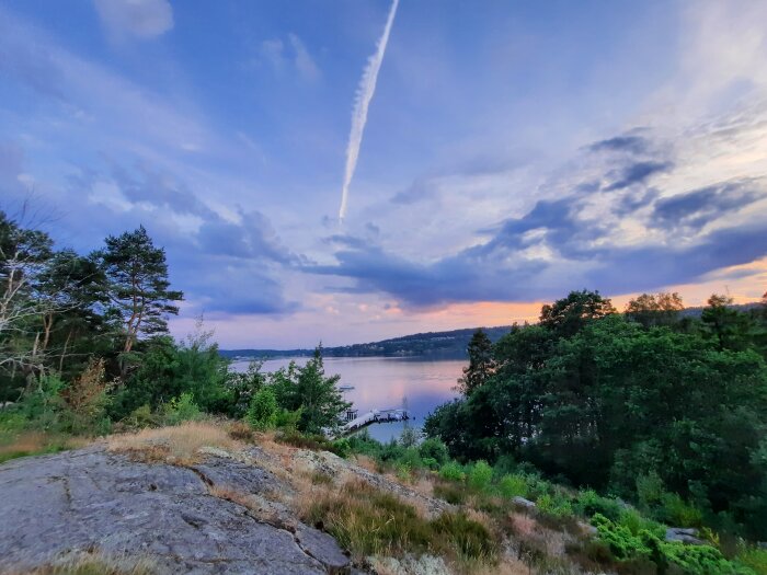 Solnedgång över vatten, träd och klippor, dramatisk himmel med kondensstråk efter flygplan.