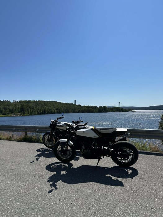 Två motorcyklar vid en sjö, med skog och en bro i bakgrunden, klarblå himmel.