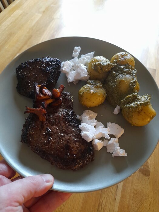 Ett måltid bestående av stekt kött, potatis med pesto och vit färskost på tallrik.