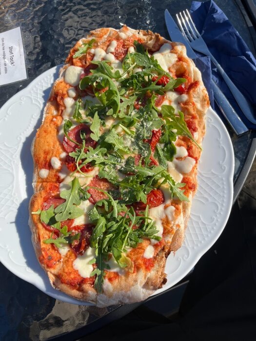 Pizza med ruccola, soltorkade tomater, salami och ost på en vit tallrik utomhus.