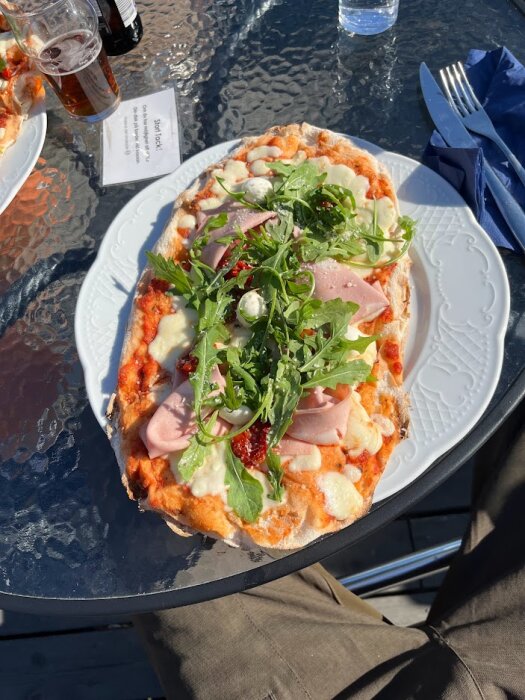 Pizza med skinka, ruccola och ost på tallrik utomhus med dryck och servis.