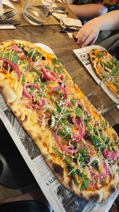 Stor pizza med rucola och rödlök på restaurang med vattenglas och meny synlig.