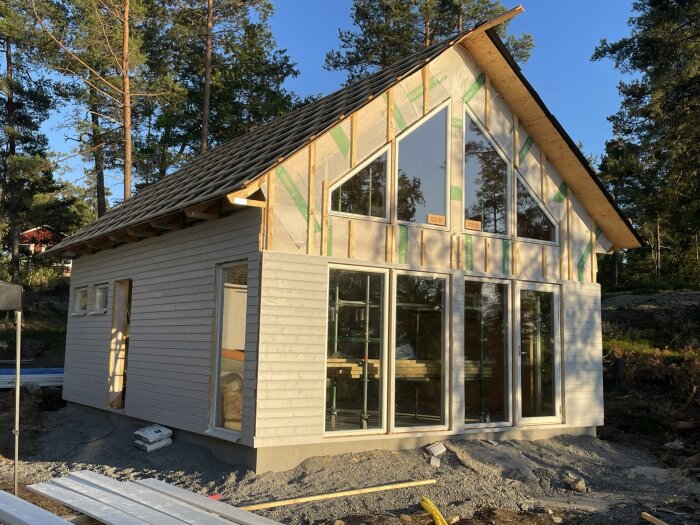 Modernt hus under konstruktion med stora fönster omgivet av skog i solnedgång.