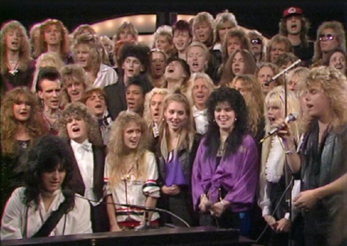 Stor grupp människor sjunger tillsammans, retrokläder, 80-tals frisyrer, mikrofoner, gemensam prestation, musikevenemang.