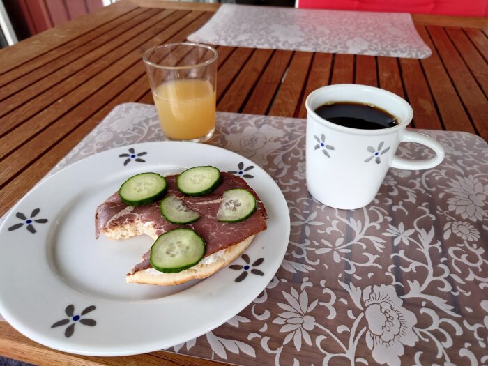 Frukost utomhus med smörgås, skinka, gurka, kaffe och juice på träbord med duk.