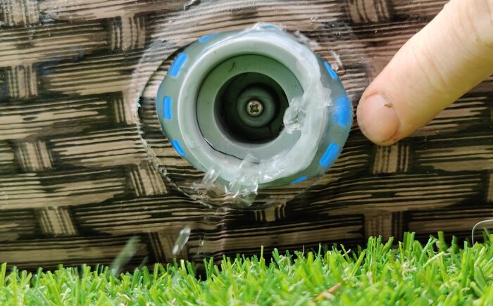 Finger vid sprinkler, klart vatten, plastfilm, träyta, konstgräs nederkant.