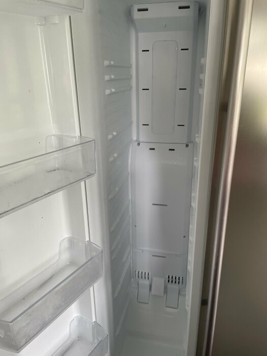 Tom kylskåpsinteriör med öppen dörr, hyllor, och genomskinliga behållare, behöver rengöring.