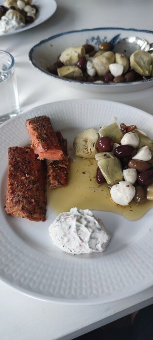 Maträtt med lax, marinerade grönsaker, mozzarella, oliver och sås, serverad på vit tallrik med vattenglas.