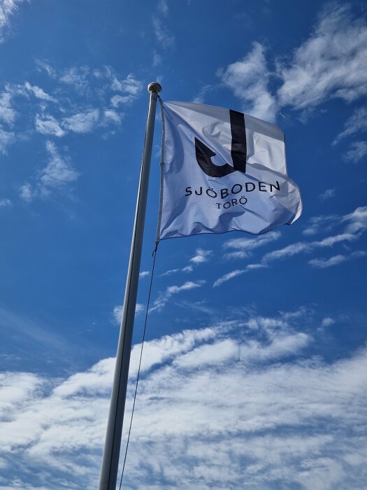 Vit flagga med svart emblem och text, upphängd på stolpe mot blå himmel med vita moln.