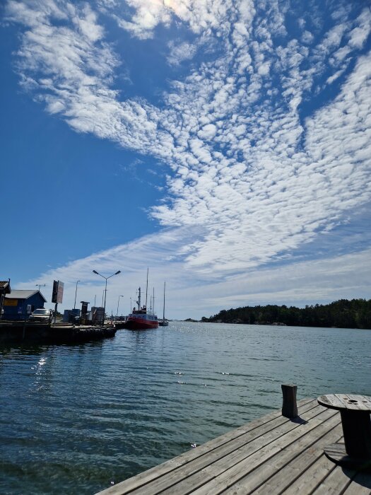 Ett träbrygga som leder till klarblått vatten, himmel med spridda moln och en röd båt vid kajen.