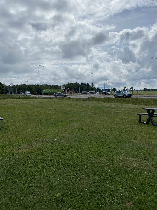 Grönt fält, picknickbord, molnig himmel, trafik i bakgrunden, gatlyktor, skyltar, ytterområde vid dagtid.