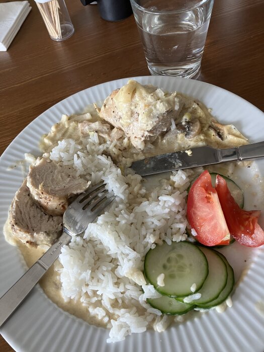 Ett måltidsarrangemang med ris, kyckling, gräddsås, tomat, gurka och ett glas vatten.