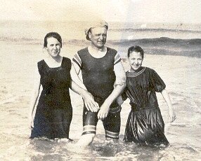 Tre personer i gammaldags badkläder står i vattnet och ler, svartvit foto, historisk känsla, stranddag.