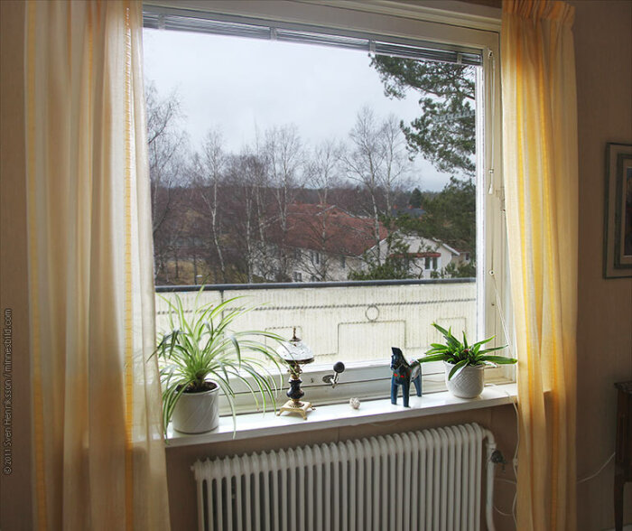 Ett fönster med öppen gardin, växter, en leksaksfigur, utsikt över träd och hus, inomhusmiljö.