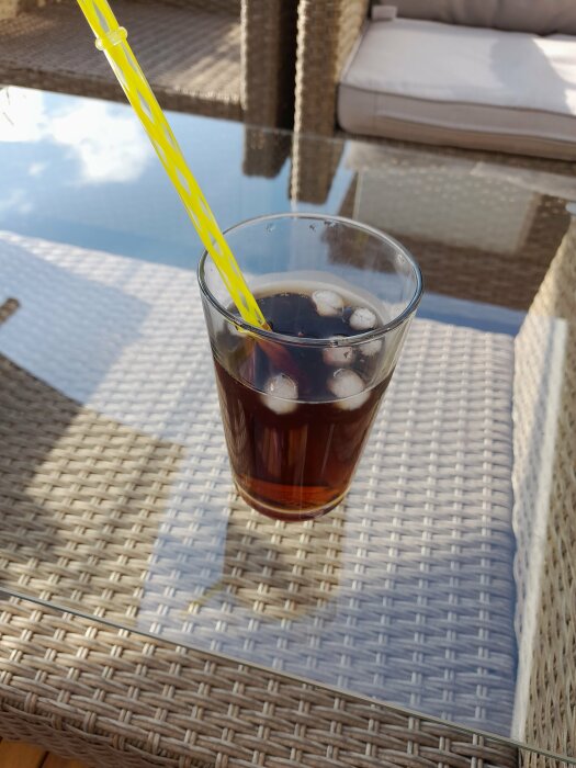 Ett glas med mörk dryck och isbitar på ett flätat bord, med ett sugrör, utomhus.