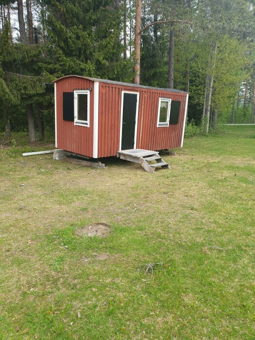Röd stuga med vita knutar, naturlig omgivning, gräs, skog, osymmetrisk trappa, ingen synlig väg.