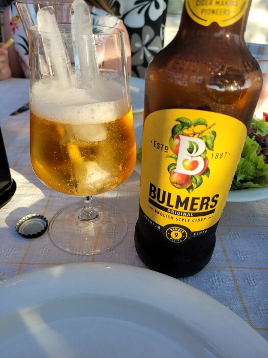 Flaska Bulmers cider, glas med is, uteservering, öppnad kapsyl, del av tallrik, sommardag.