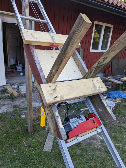 Improviserad arbetsstation utomhus med trappstegar, träklossar, byggmaterial, verktyg och röd byggnad i bakgrunden.