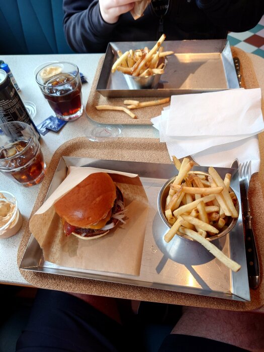 Person äter hamburgare och pommes frites vid bord med drycker och servetter.