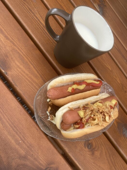 Två varmkorvar med senap och ketchup, rostad lök, mugg med mjölk, träbord.