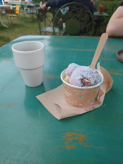 Glass i bägare på grönt bord, träsked, vit mugg, personer i bakgrunden, utomhusmiljö.