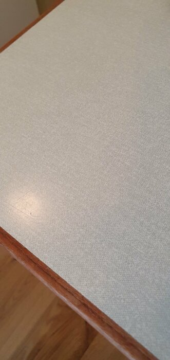 Ett bord hörn med sliten träkant och beige textilerad yta.