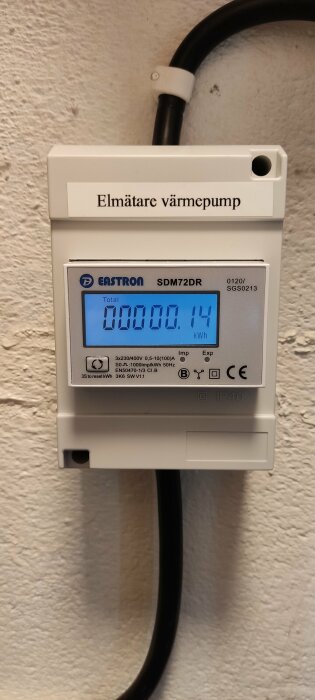 Elmätare med digital display visar energiförbrukning för värmepump, fast monterad på vit vägg.