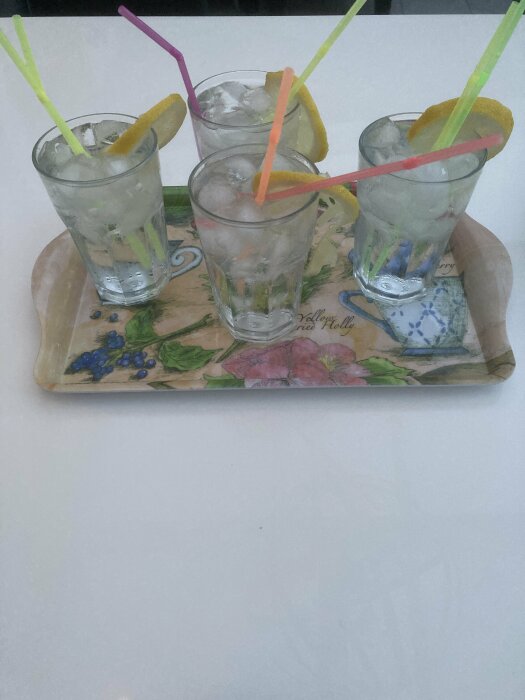 Glass med is, vatten, sugrör och citronskivor på ett bricka med blommönster.