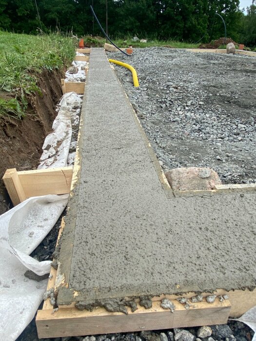 Nygrävt dike med betongvägg, träformar, grusunderlag och gul slang ute på ett anläggningsområde.