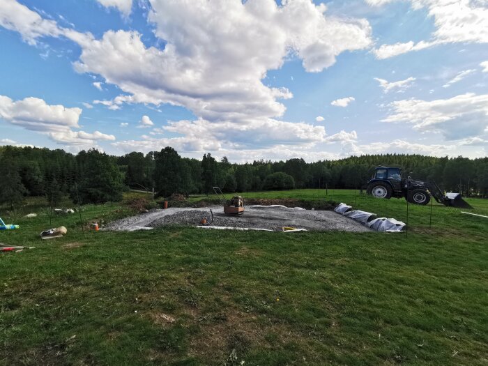 Åker med anlagd damm, traktor, skogsbakgrund, molnig himmel, pågående markarbete.