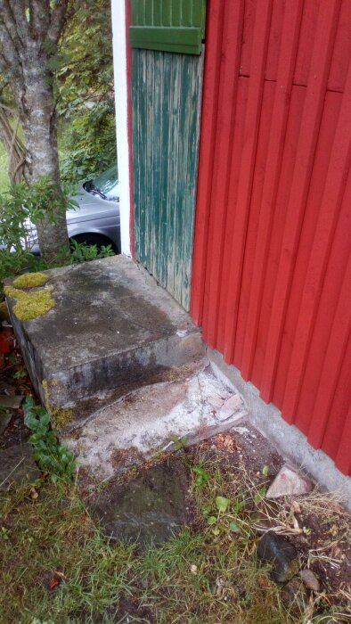 En sliten trätrappa, mossigt betongsteg, del av röd vägg och en grön dörr.