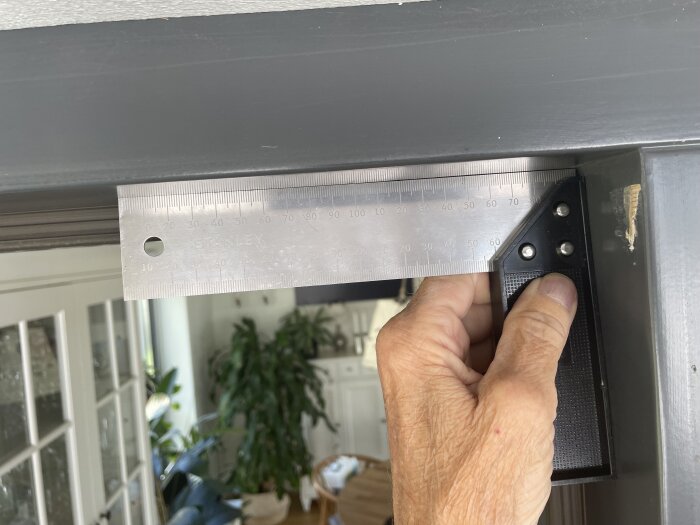 En hand håller en metallisk linjal mot en dörrkarm för att mäta eller kontrollera räthet.