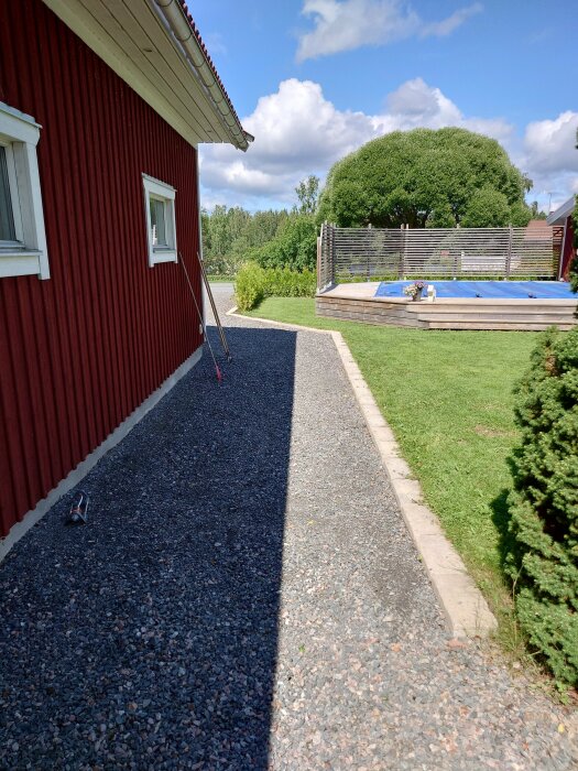 Röd stuga, grusgång, grönt gräs, trädäck med pool, ljust väder, en ensam sko.