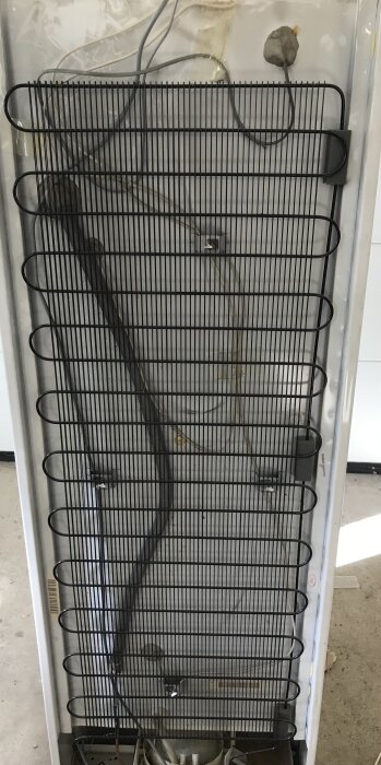 Bakdel av öppen kyl, svart kondensatornät, kablar, termostat, oorganiserade delar, behöver städning.