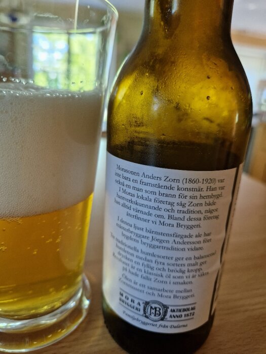 Glas öl, flaska med etikett, text om Anders Zorn, Mora Bryggeri, bordsyta.