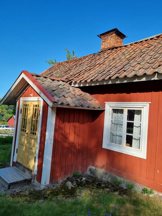 Rött trähus med tegeltak, vit fönsterkarm, gul dörr, frodig natur, traditionell skandinavisk arkitektur, sommartid.