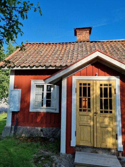 Ett traditionellt rött svenskt trähus med en gulfärgad dörr och vita fönster under en klarblå himmel.