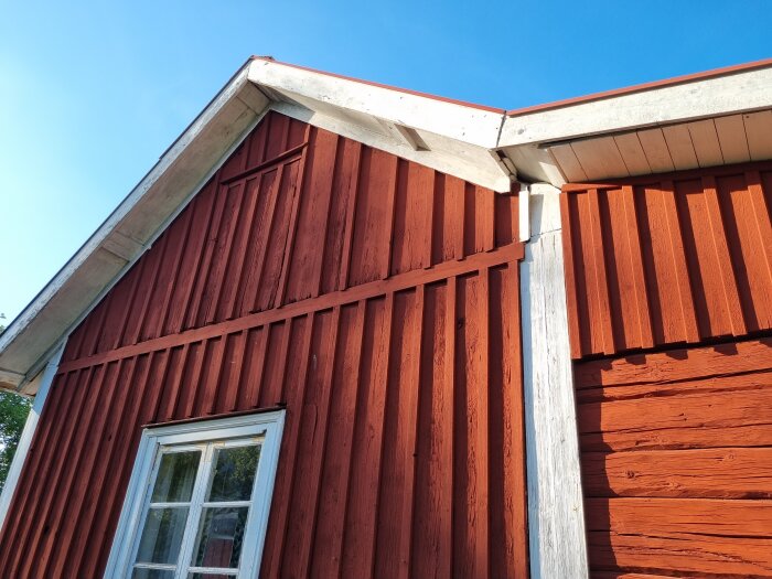 Faluröd timmerstuga, vitkantad takfot, soligt, fönster, traditionell svensk landsbygdsarkitektur.