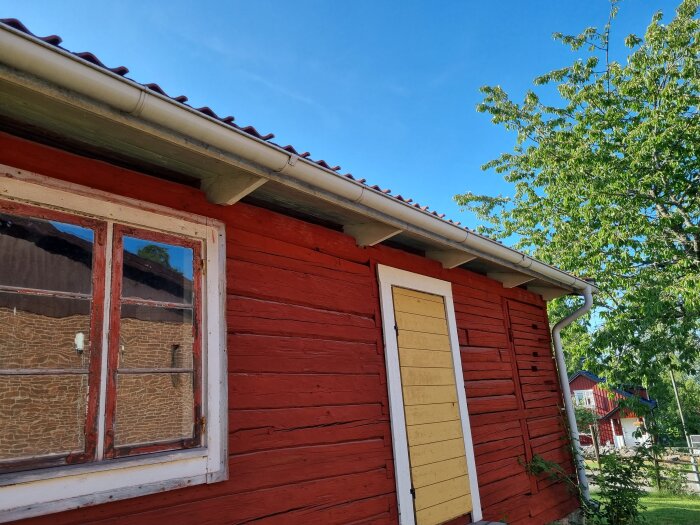 Rödmålad trästuga med vita knutar, gult fönsterfoder, grön natur och blå himmel.