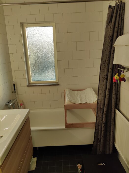 Badrum med vita kakelväggar, badkar, duschdraperi, handfat och trähylla. Frostat fönster släpper in ljus.