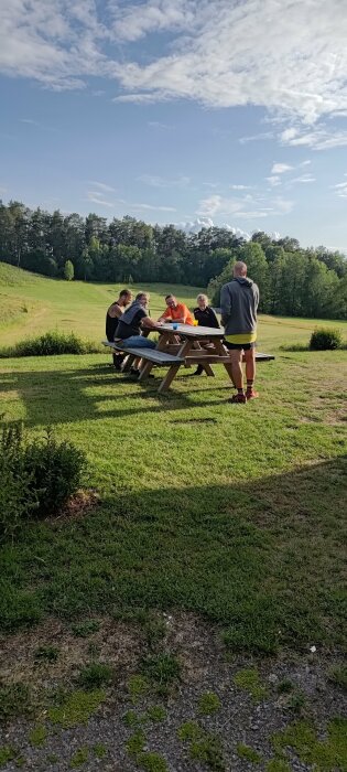 Grupp människor samlade runt picknickbord i grönt utomhuslandskap med blå himmel och moln.